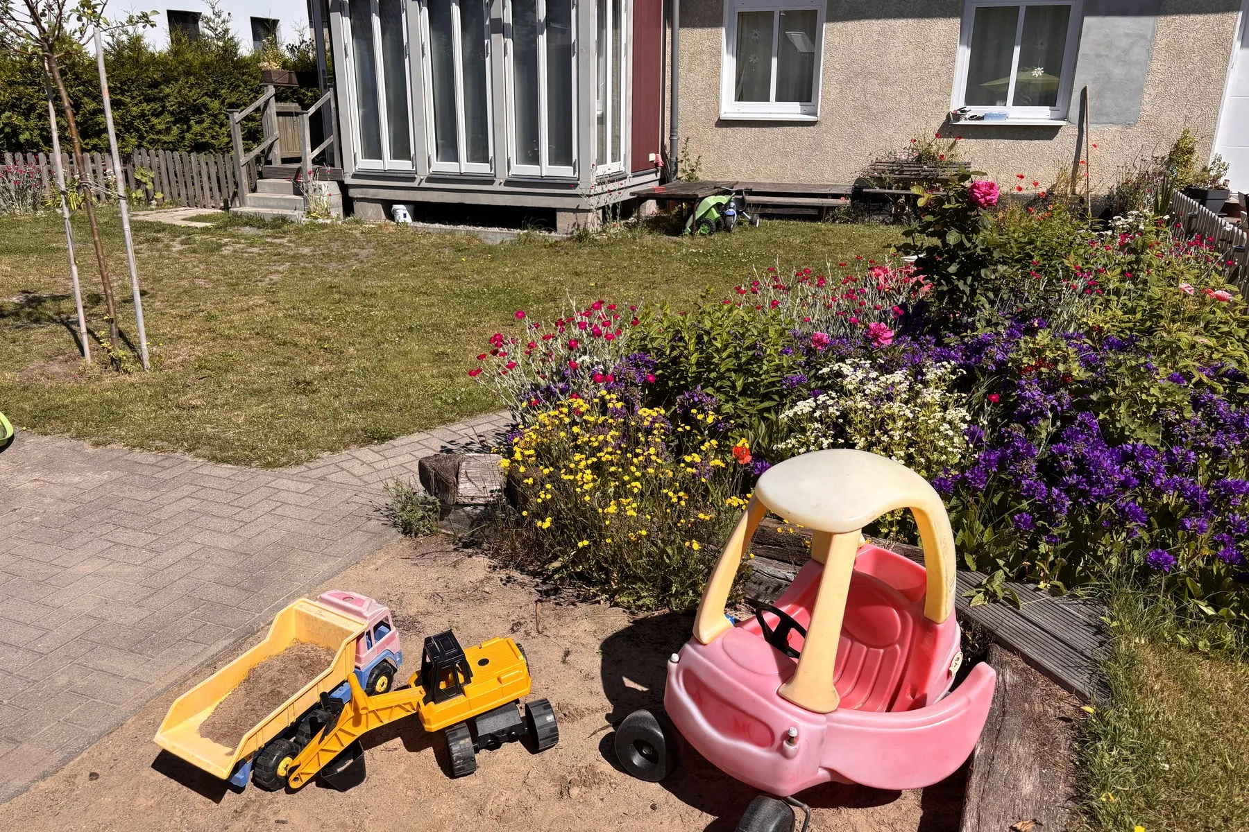 Gepflasterter Spielbereich mit Kipplaster und Rutscheauto vor einem Blumenbeet