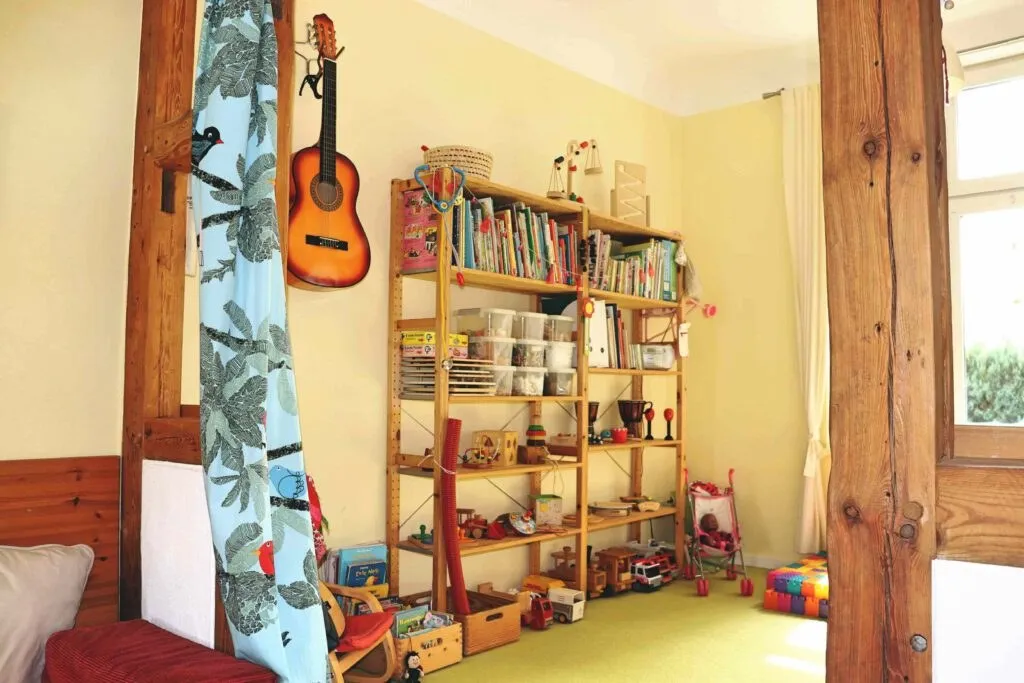Spielraum mit Fachwerkbalken: an der Wand hängt eine Gitarre, daneben ein hohes Regal mit Bilderbüchern, Trommeln und Holzspielzeug
